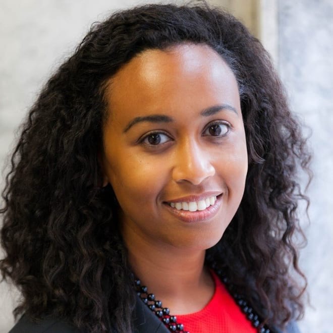 A smiling Black woman with long curly dark hair wearing a red top and black jacket