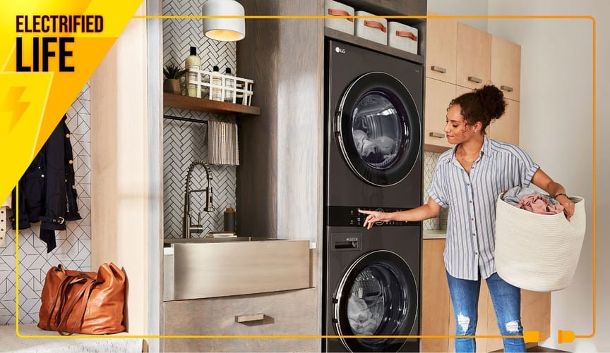 Woman with brown curly hair stands with load of laundry in front of gleaming metal stacked clothes dryer and washer..