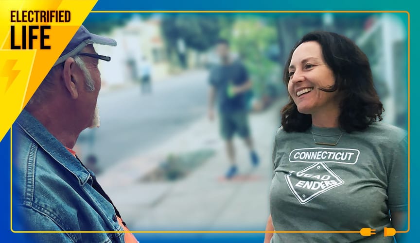 Woman with dark hair talks to man with white sideburns and beard on the sidewalk.