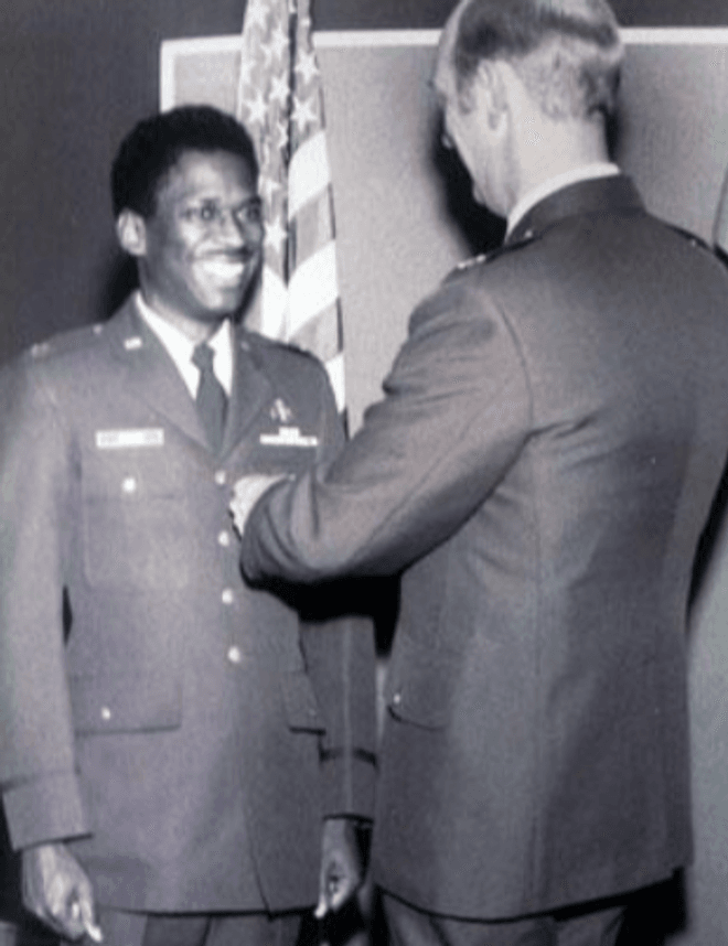 Two men in uniform. A taller white man pins a medal on the uniform of a smiling Black man. An American flag in the background