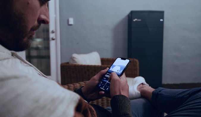 Man holding smartphone displaying Lunar Energy app, with Lunar Energy battery/inverter in the background