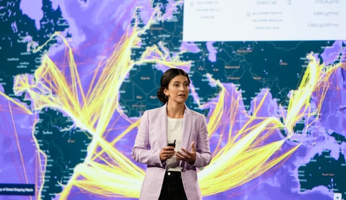 A woman with dark hair and wearing a lavender jacket stands in front of a map of the world's ocean cargo routes