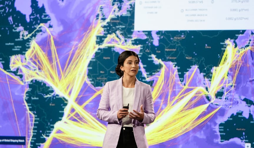 A woman with dark hair and wearing a lavender jacket stands in front of a map of the world's ocean cargo routes