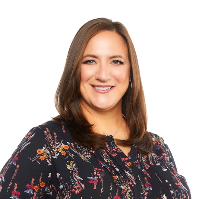 A smiling woman with medium skin tone and shoulder length brown hair wearing a multicolored shirt