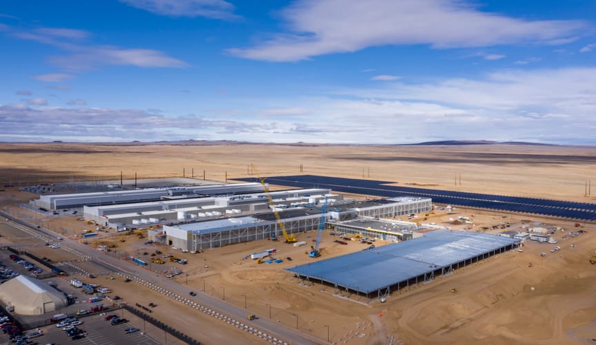 A large complex of elongated industrial buildings next to a large solar array in a vast desert landscape under a blue sky