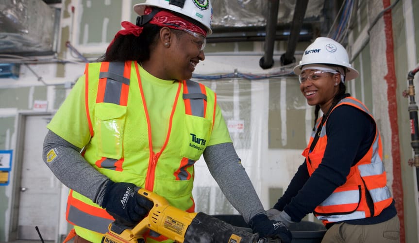 Two women with medium brown skin wearing hard hats and safety gear smile while working on an indoor construction site