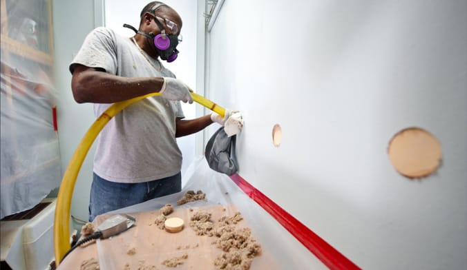 Black man holds yellow tube against hole in wall inside home.