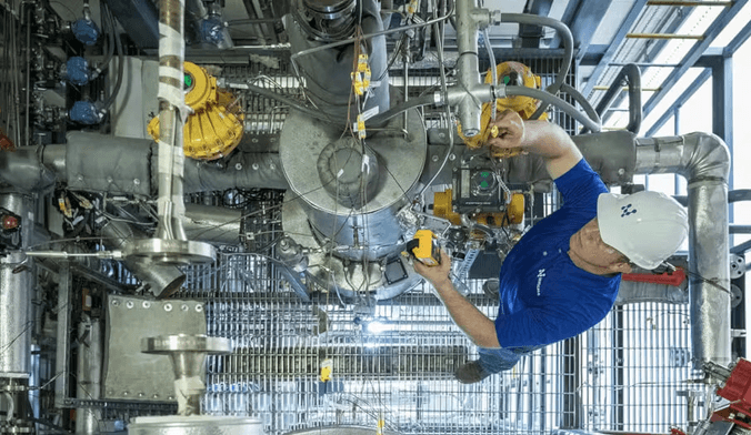 A worker in a white hard hat and blue polo shirt works on a complex piece of equipment