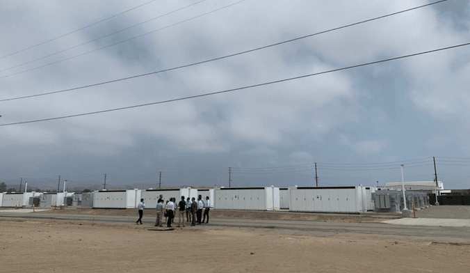 A crowd of  people gathered near utility-scale battery plant consisting of long white metal enclosures