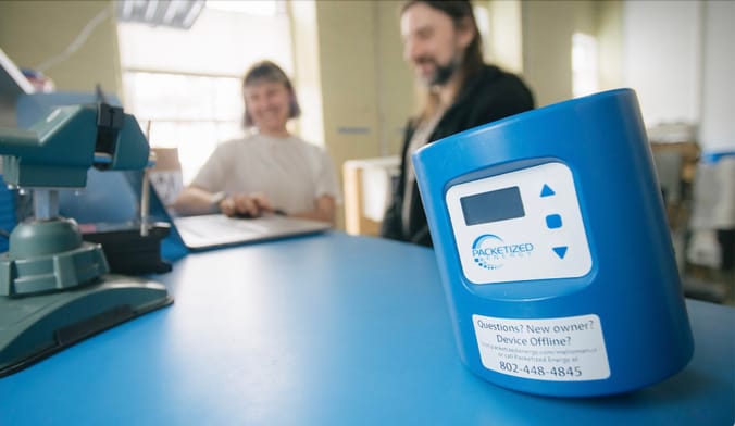 A blue Packetized Energy smart control device sits on a lab bench with two people in the background