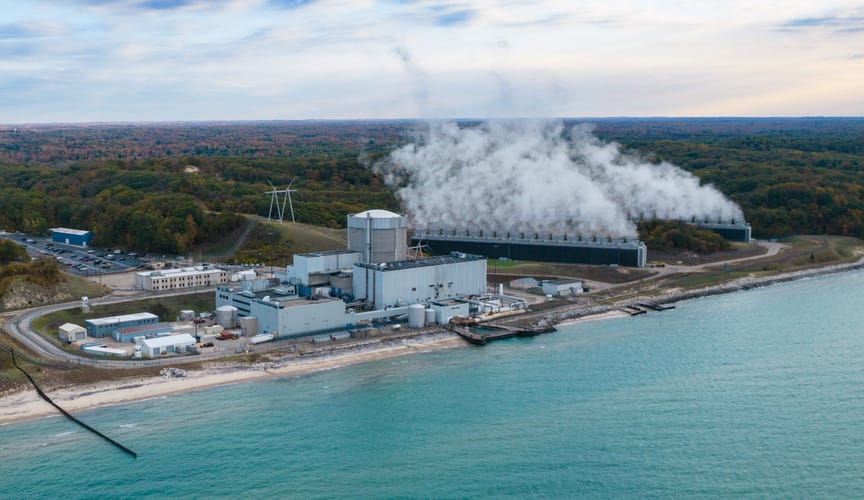 A large industrial facility emitting large clouds of steam sits on the back of a vibrant blue lake
