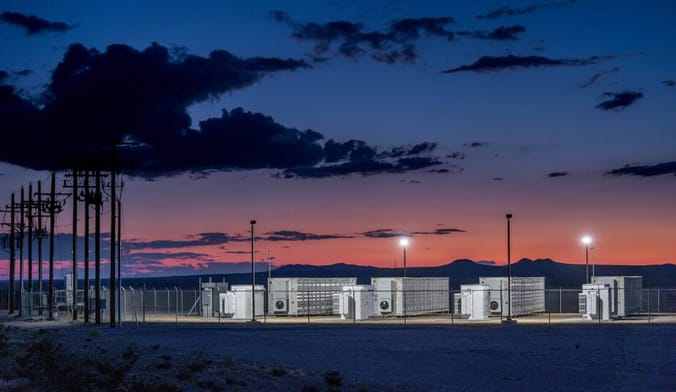 A row of large white rectangular metal battery enclosures against the backdrop of a dramatic pink and purple sunset