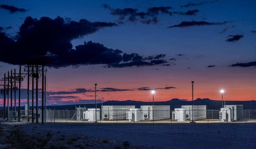 A row of large white rectangular metal battery enclosures against the backdrop of a dramatic pink and purple sunset