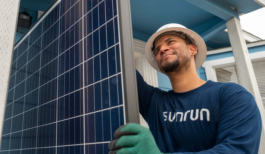 A man wearing a white hard hat and green gloves holds a solar panel