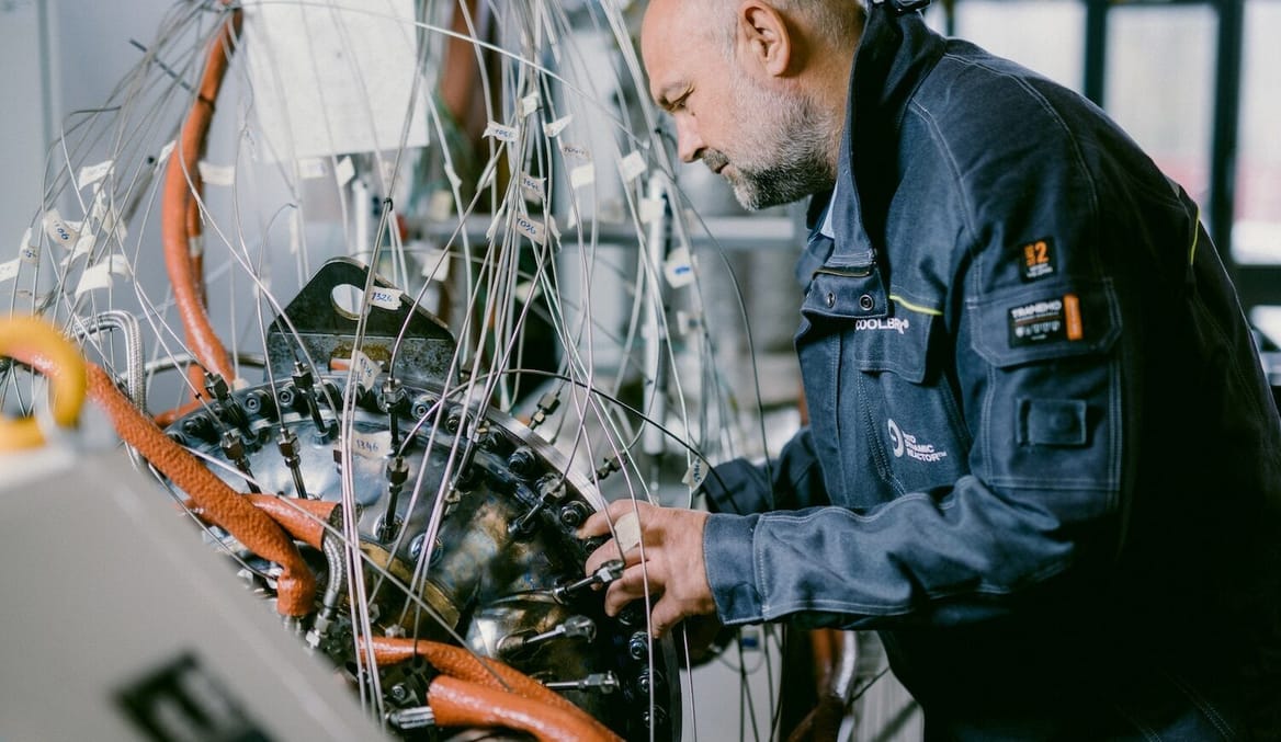 A man with light-toned skin and gray hair and beard wearing safety clothes looks closely at some laboratory equipment