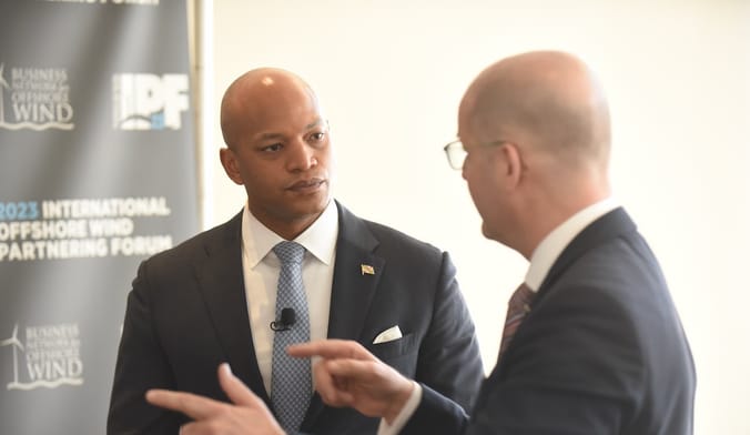 Two men have a conversation, with a poster advertising an offshore wind conference behind them.