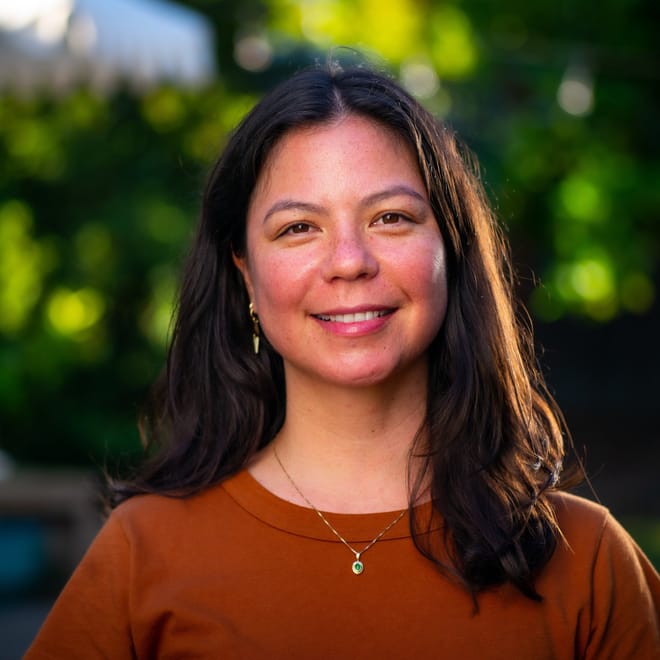 A smiling woman with medium skin and shoulder-length dark hair wearing a dark orange top