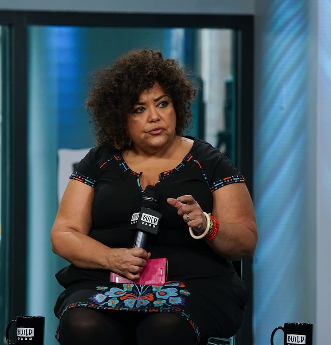 A woman with black curly hair wearing a black dress is seated and speaking into a microphone