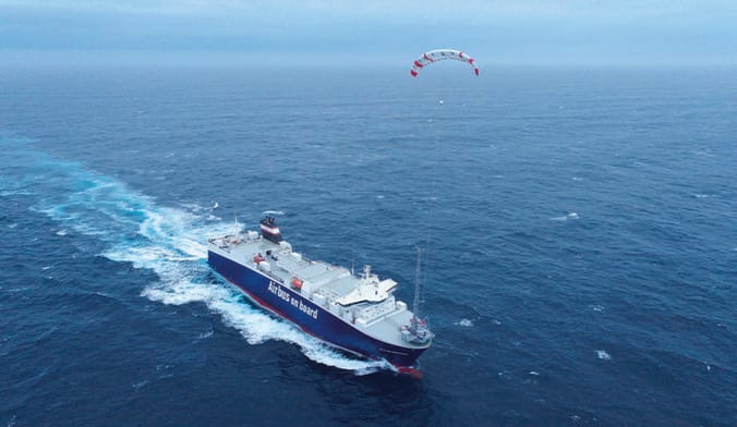 A large ship sails across the ocean with a tethered, rainbow-shaped white kite flying above it