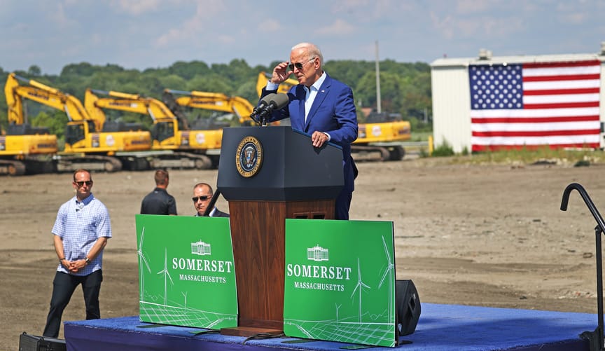 A man with white hair wearing a blue jacket speaks on a dais outdoors, flanked by a row of bulldozers and a large US flag