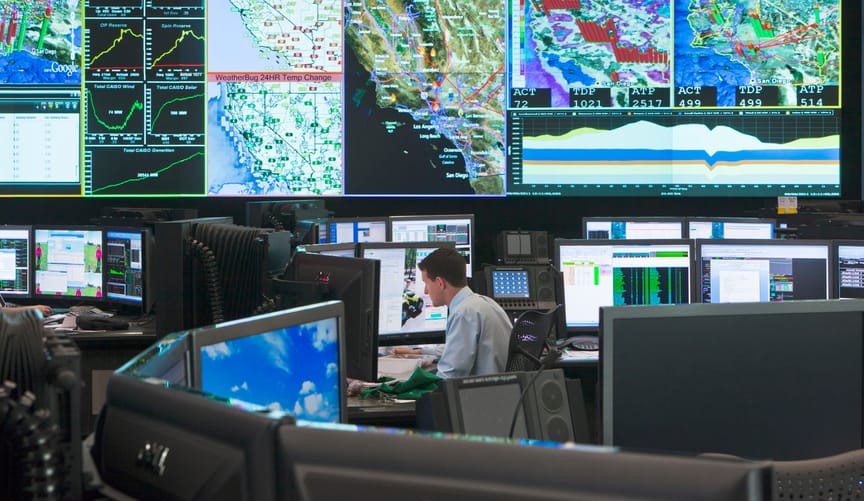 A man in a white shirts sits in a utility control room filled with monitors and wall displays