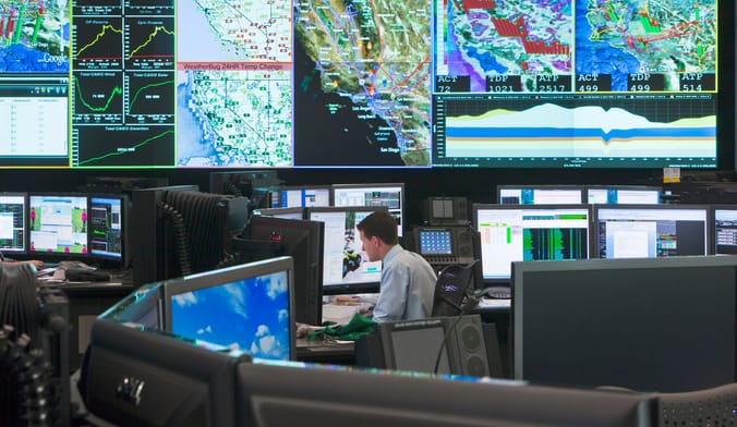 A man in a white shirts sits in a utility control room filled with monitors and wall displays