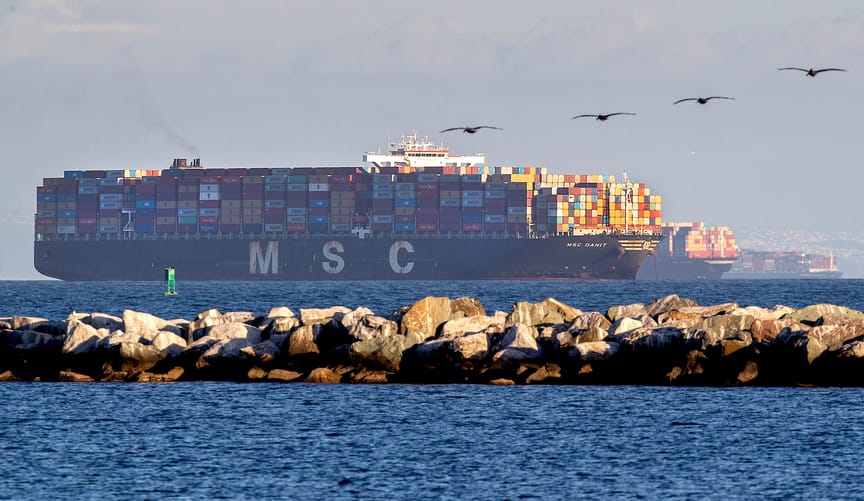 a large cargo ship piled high with colorful shipping containers sits off the coast of Long Beach