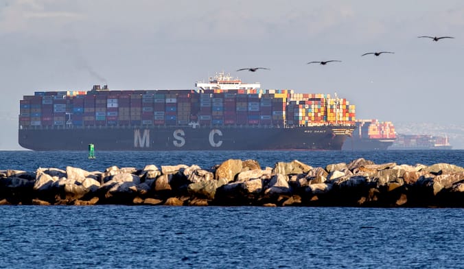 a large cargo ship piled high with colorful shipping containers sits off the coast of Long Beach