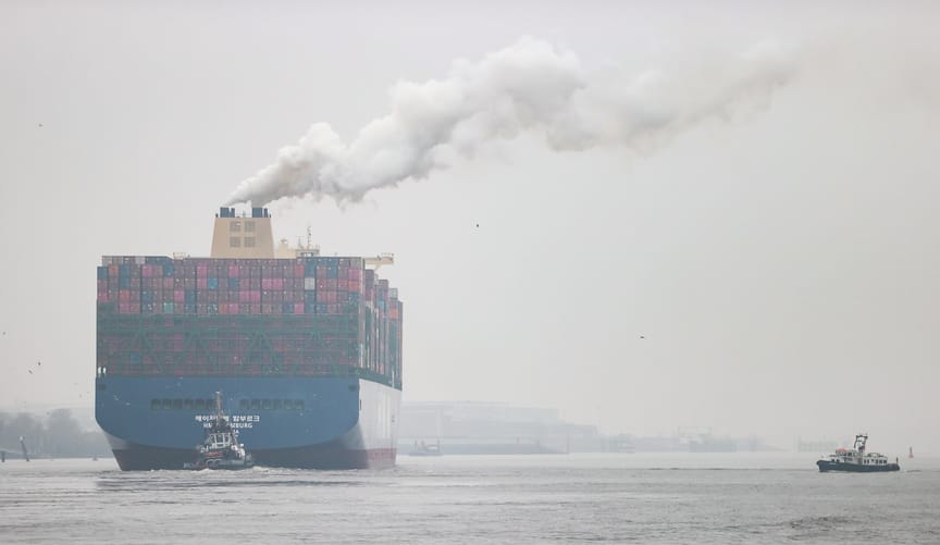 A very large cargo ship sails at sea with a plume of white smoke rising from it