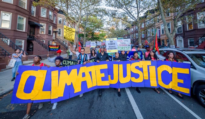 A large group of people walk through a neighborhood lined with brownstone houses holding a sign that says climate justice