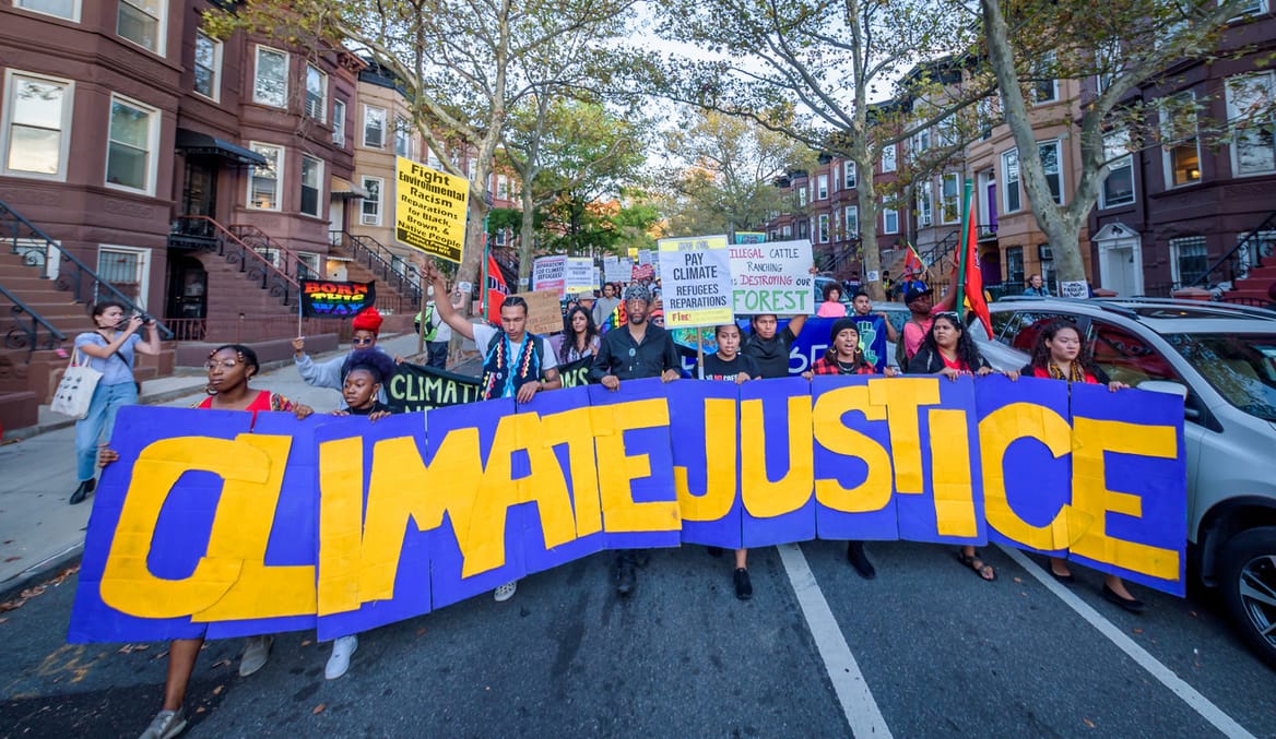 A large group of people walk through a neighborhood lined with brownstone houses holding a sign that says climate justice