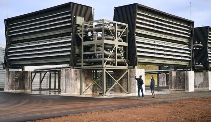 Two men in coats stand in front of a large industrial plant with metal machinery to remove carbon from the air