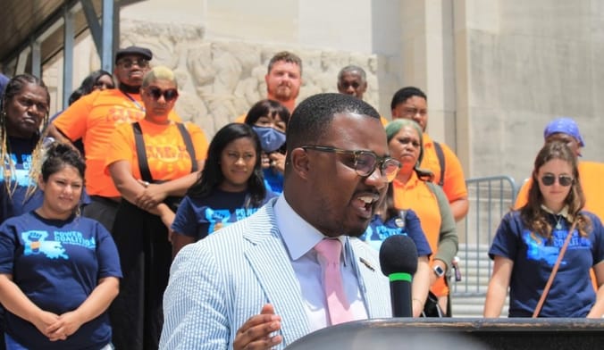 A Black man wearing glasses and a blue seersucker suits speaks into a microphone at an outdoor event with people behind him