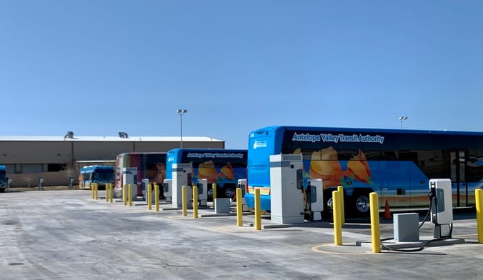 A large bus depot where several electric buses are charging under a cloudless blue sky