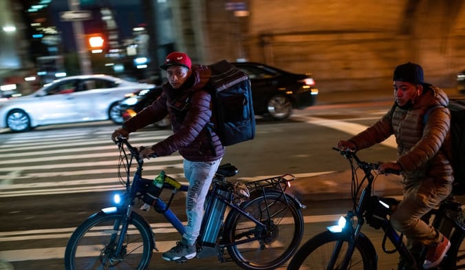 Two men on e-bikes with delivery courier backpacks ride among car traffic on the streets of Manhattan at night