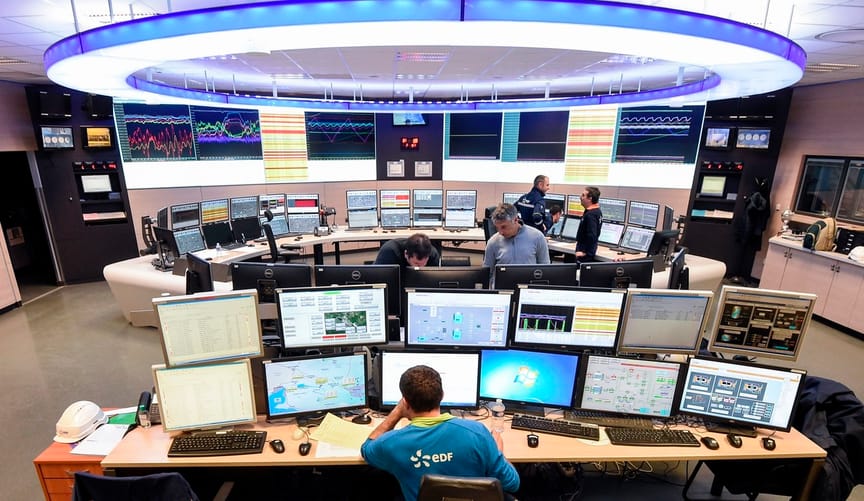 A man sits in front of a large bank of computer monitors in a utility control room