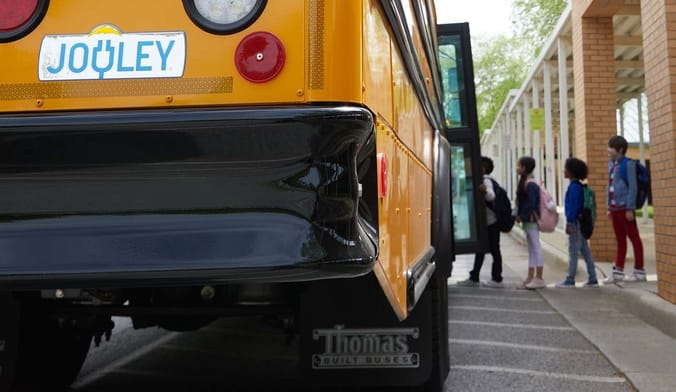 Children line up in front of a school to get on a waiting yellow school bus
