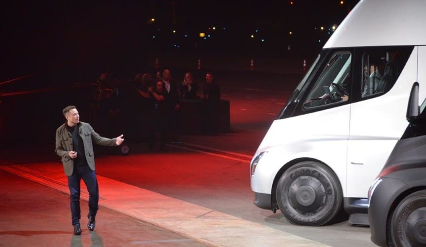 A man in a dark jacket and black pants gestures at a large white vehicle on stage at an event