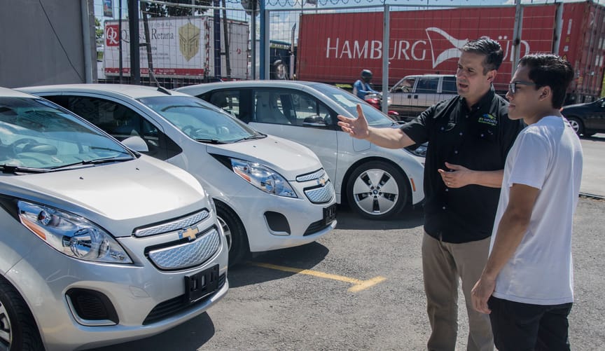 Two men stand in a car dealership lot looking at electric vehicles