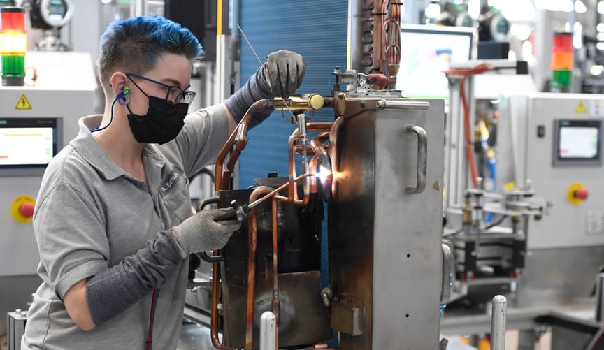 A factory worker with blue hair and eyeglasses wearing a gray shirt uses a soldering gun on a heat pump