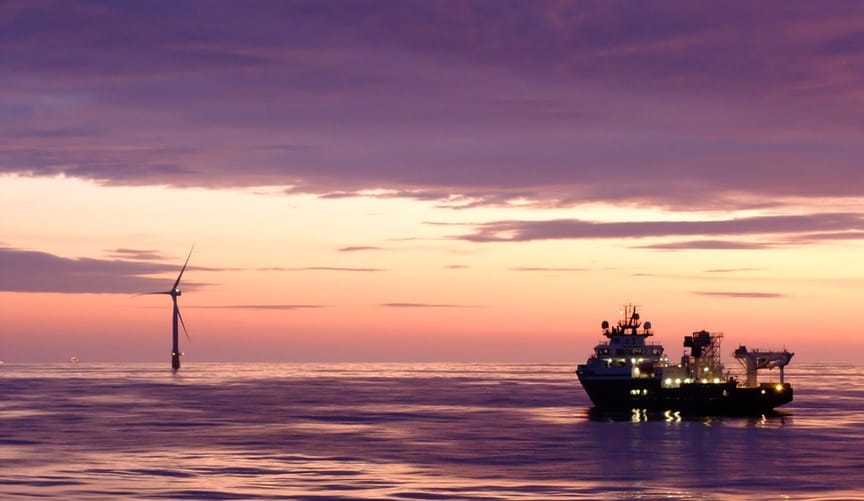 A wind turbine and a ship in the ocean against the backdrop of a dramatic pink and purple sunset