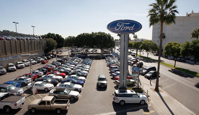 Dozens of cars, trucks and minivans are parked in an outdoor dealership lot with a large Ford sign