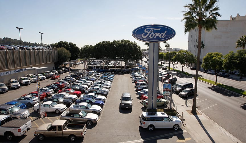 Dozens of cars, trucks and minivans are parked in an outdoor dealership lot with a large Ford sign