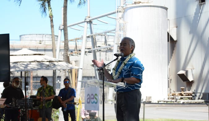 A man with gray hair wearing a lei speaks to a crowd from a lectern