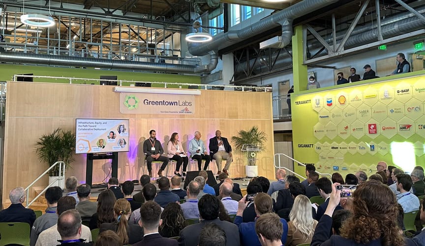 four speakers sit on a dais in a large conference room in front of an audience