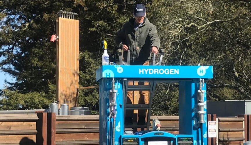 A worker in a green coat stands on a ladder servicing a blue hydrogen fuel pump in a wooded area