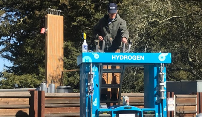 A worker in a green coat stands on a ladder servicing a blue hydrogen fuel pump in a wooded area