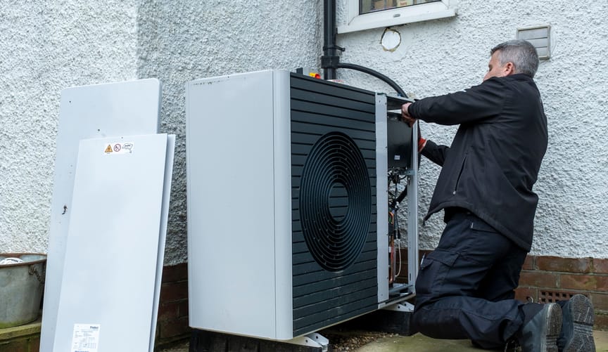 A repairman kneels while performing maintenance on a gray heat pump