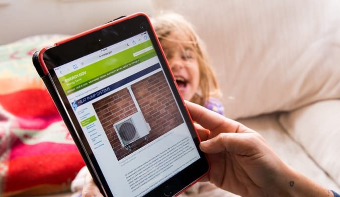 A woman reads about heat pumps on a red tablet while her young daughter laughs on the couch