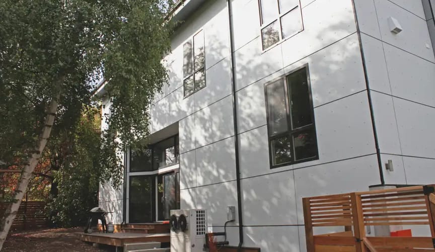 A multistory white home with trees in the yard and a large heat pump